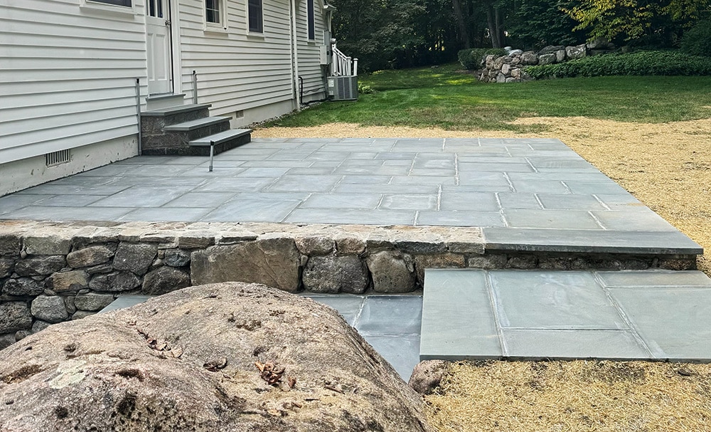 Stone patio with steps leading to a white house. A large rock is in the foreground, and a grassy yard is in the background.