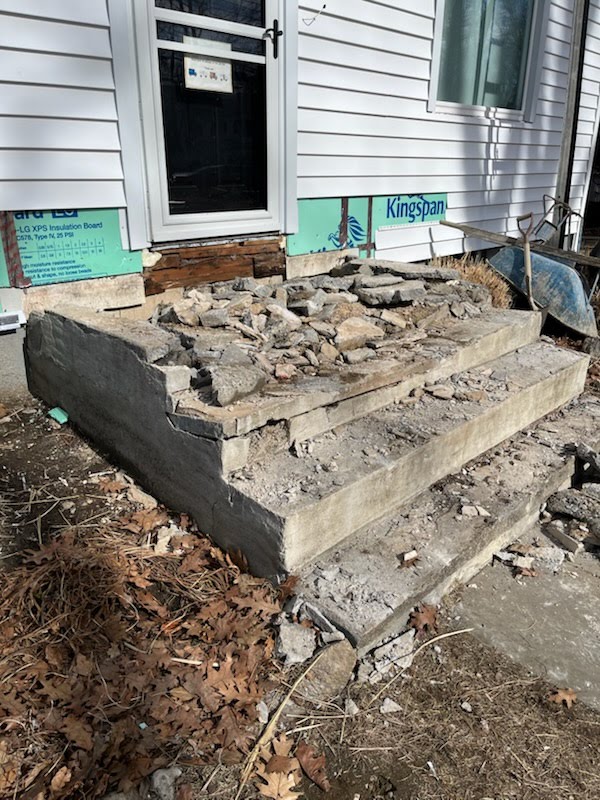 Damaged concrete steps with broken and loose pieces in front of a house under partial renovation, with construction materials nearby.