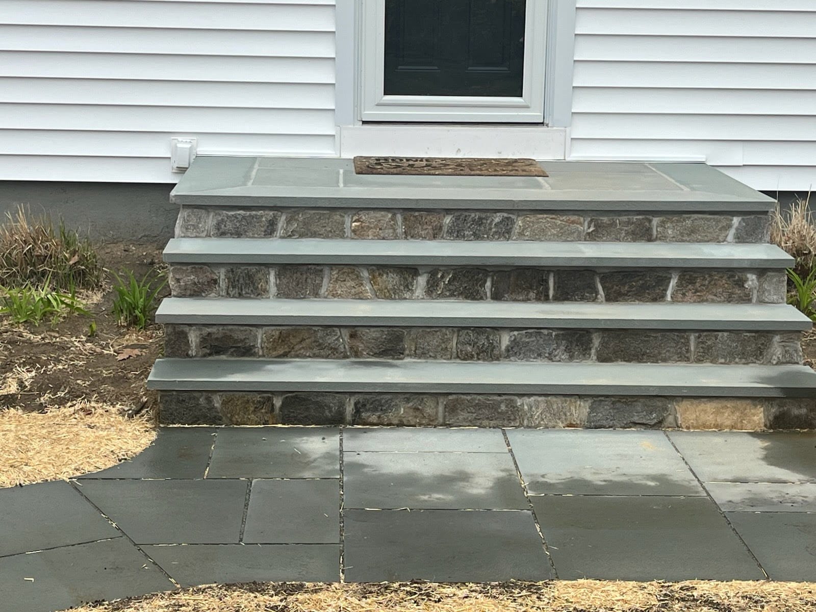 Stone steps with a doormat lead up to a dark door on a white paneled house, with a stone pathway and some straw-covered ground in front.