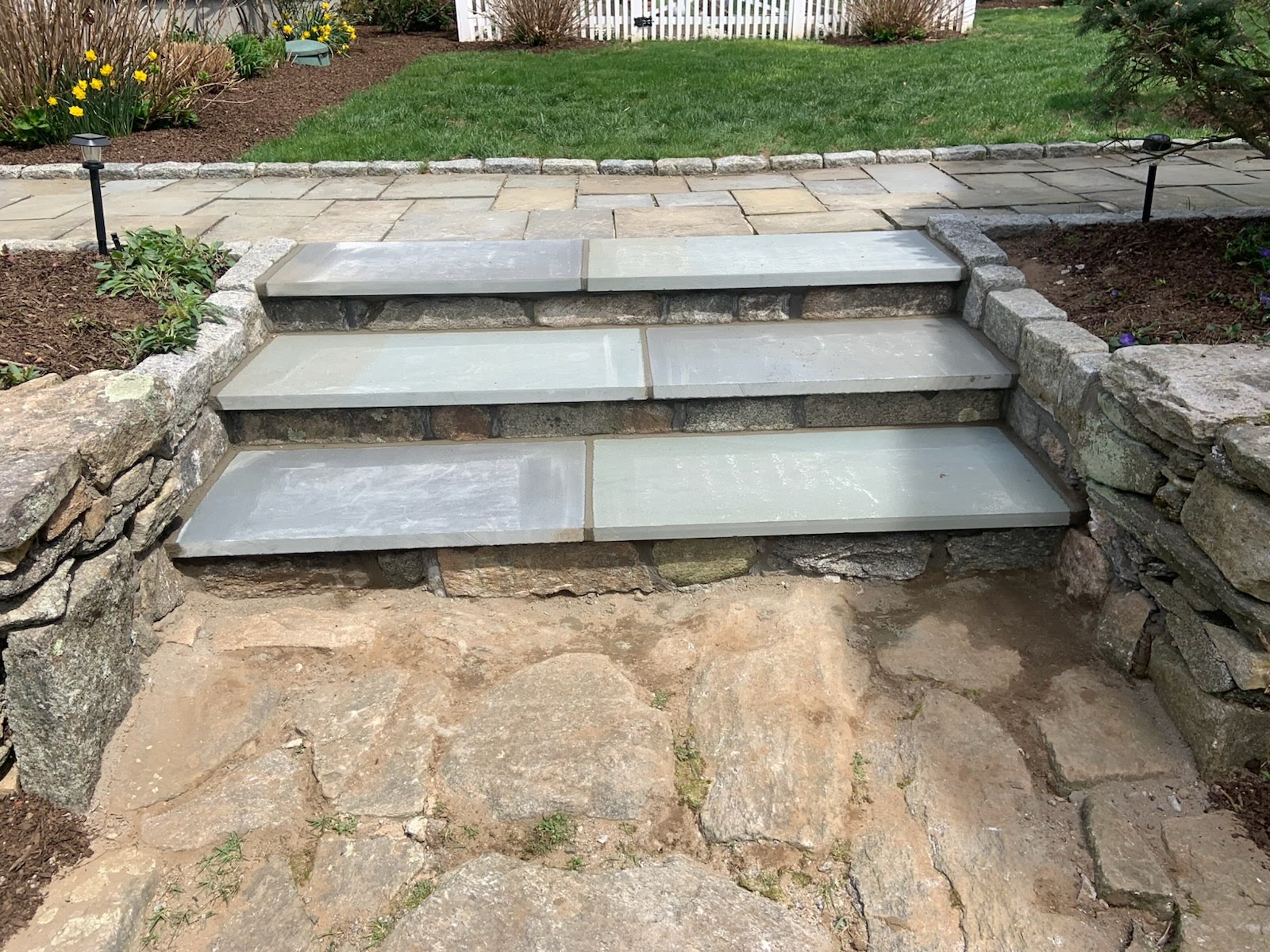 Three stone steps with wide risers lead up from a flagstone path, bordered by stone retaining walls and surrounded by garden beds.