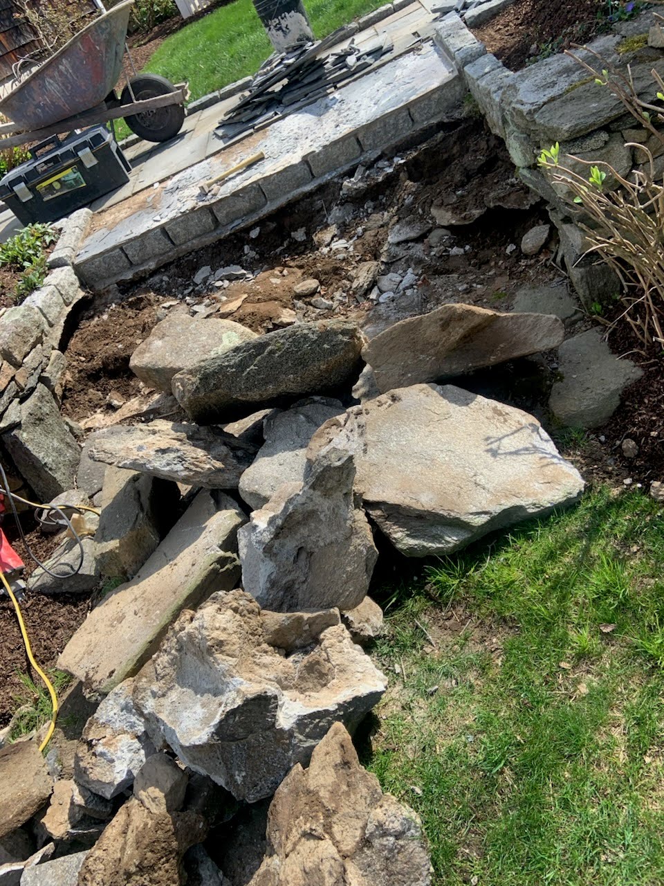 Large broken stones and rubble are scattered on the ground near a partially dismantled stone wall and patio area in a garden setting.